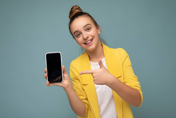 Photo of beautiful smiling teenager good looking wearing casual stylish outfit standing isolated on background with copy space holding smartphone showing phone in hand with empty screen display for - Fotoğraf, Görsel