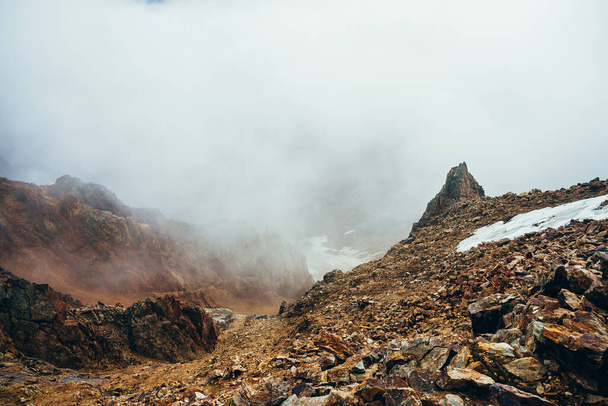 Όμορφος μυτερός γκρεμός στην ψηλή βραχώδη άκρη του βουνού με χιόνι ανάμεσα σε πυκνά χαμηλά σύννεφα. Ατμοσφαιρικό μινιμαλιστικό αλπικό τοπίο Σημαδεύει μια πέτρα κοντά στην άβυσσο σε ένα μεγάλο σύννεφο. Υπέροχο ορεινό τοπίο. - Φωτογραφία, εικόνα