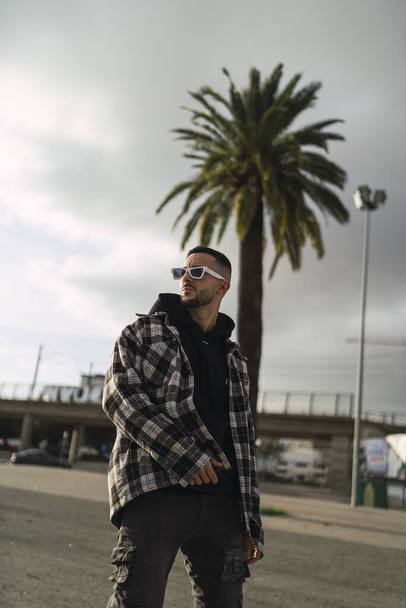 A vertical shot of a stylish guy wearing a plaid jacket, a black hoodie, black pants, and sunglasses - Fotoğraf, Görsel