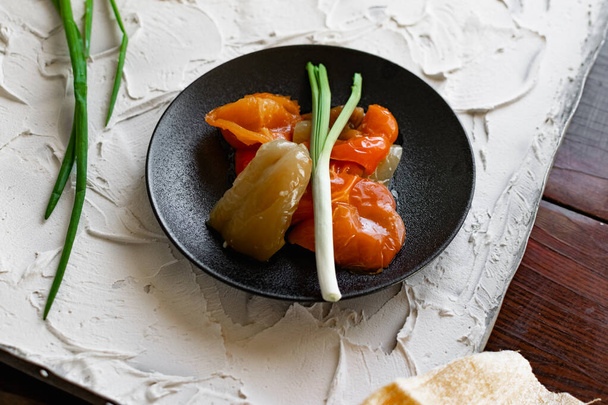 Pickled sweet bell pepper, cut into slices, multicolored vegetables in oil on a black matte plate and a stone white background. The dish is decorated with green leeks - Foto, imagen