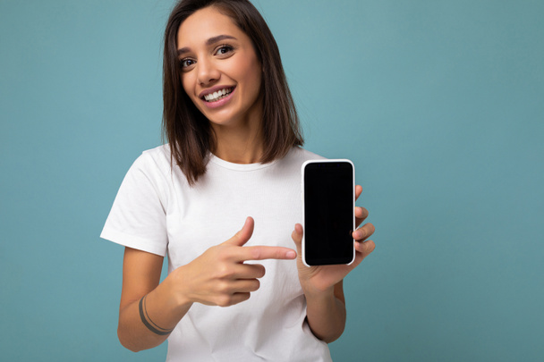 Фото красивой улыбающейся молодой женщины, красивой в повседневной стильной одежде, стоящей изолированно на заднем плане с копировальным пространством со смартфоном, показывающим телефон в руке с пустым дисплеем экрана для - Фото, изображение