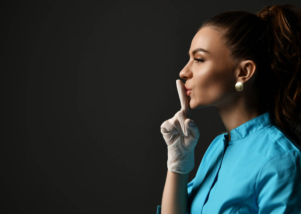 Young woman doctor in blue medical gown and latex gloves holds finger at her lips gesturing shh sign calling for silence - Foto, Imagen