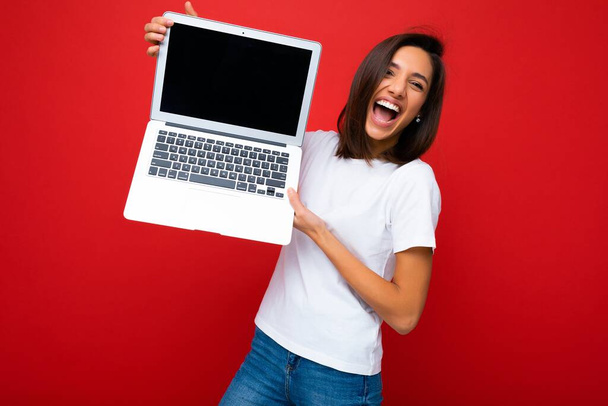 Krásná šťastná radostná mladá žena s krátkým tmavým brunet účes drží počítač notebook při pohledu na kameru na sobě bílé tričko a džíny izolované přes červenou zeď pozadí - Fotografie, Obrázek