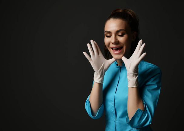 Young brunette woman doctor nurse in blue medical gown and latex gloves holds hands at face happy screaming - Fotó, kép