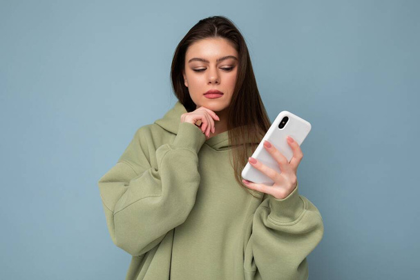 Photo of serious thoughtful Beautiful young brunette woman wearing stylish green hoodie using mobile phone writing message isolated on background looking at smartphone display - Photo, Image
