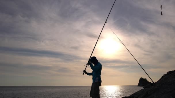 Fisherman Throws a Fishing Rod Pole Into the Sea Standing on the Rocks Slow Motion Silhouette of fisherman on the rocks seacoast at Phuket thailand in Beautiful sunset or sunrise time - Footage, Video