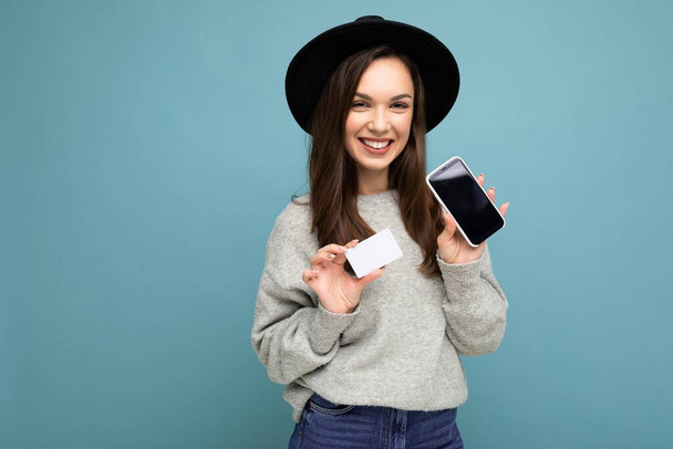 Piękny uśmiech szczęśliwy młoda brunetka kobieta noszenie czarny kapelusz i szary sweter izolowane na niebieskim tle trzymając kartę kredytową i telefon komórkowy z pustym wyświetlaczem do makiety patrząc w aparat - Zdjęcie, obraz