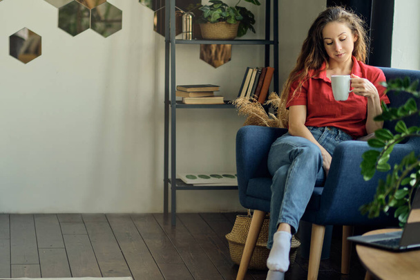fiatal nő egy csésze kávéval a kezében ül egy székben az ablak mellett egy hangulatos nappaliban - Fotó, kép