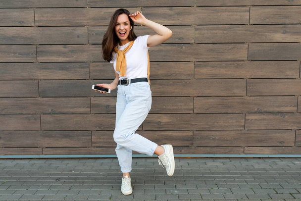 Full length body size photo of pretty brunet young happy woman wearing casual clothes and listening to music via wireless earphones standing in the street holding and using mobile phone looking at - Foto, Imagen