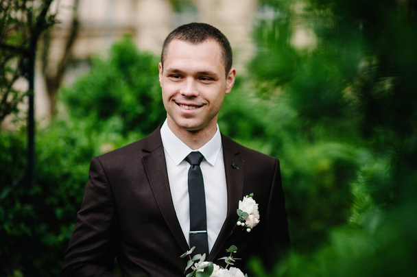 The attractive groom in a suit and tie wiht boutonniere or buttonhole on the jacket, is stands on the background greenery in the garden, park. Nature. - Фото, изображение