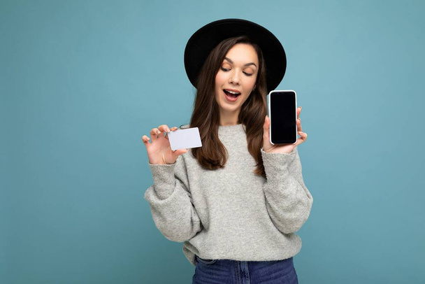 Fénykép vonzó pozitív fiatal barna nő visel fekete kalap és szürke pulóver elszigetelt kék háttér gazdaság hitelkártya és mobiltelefon üres kijelző a mockup keres a - Fotó, kép