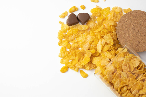 fresh crispy cornflakes in a glass jar stand on the table, pralines of chocolate lie next to, on a white background - Photo, Image