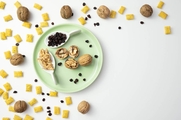raisins and cornflakes are scattered on the table, near walnuts on a green plate, on a white background - Photo, Image