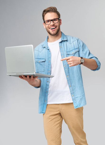 Concentrated young bearded man wearing glasses dressed in jeans shirt holding laptop isolated over grey studio background - Foto, afbeelding