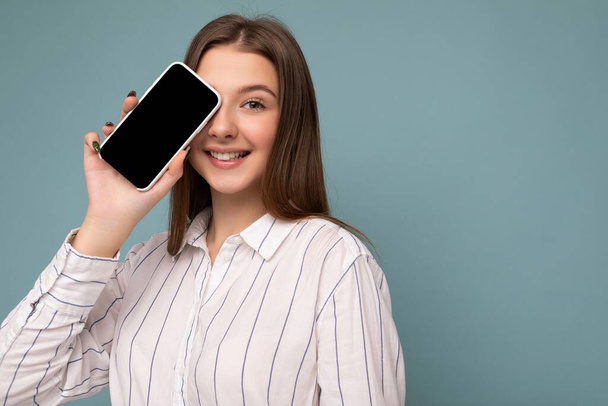 Крупним планом фотографія позитивної привабливої молодої блондинки в повсякденній білій сорочці ізольовані на синій стіні, що тримає смартфон і показує мобільний телефон з порожнім екраном для вирізання
 - Фото, зображення