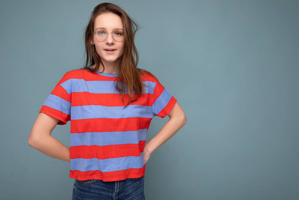 Pěkně plachá mladá studentka stojící izolovaně na modrém pozadí zdi drží ruce za sebou při pohledu na kameru nosí pruhované tričko a optické brýle - Fotografie, Obrázek