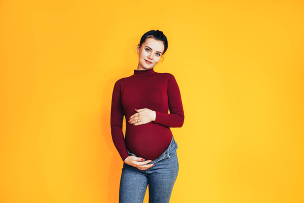 Una mujer embarazada con un suéter rojo y vaqueros azules sobre un fondo amarillo. El cuidado de la salud de las mujeres embarazadas. - Foto, imagen
