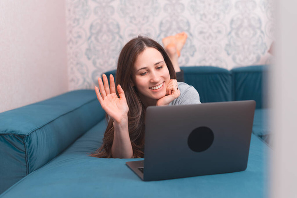Фото молодой женщины, улыбающейся и разговаривающей с друзьями в Интернете на ноутбуке, лежащей на диване - Фото, изображение