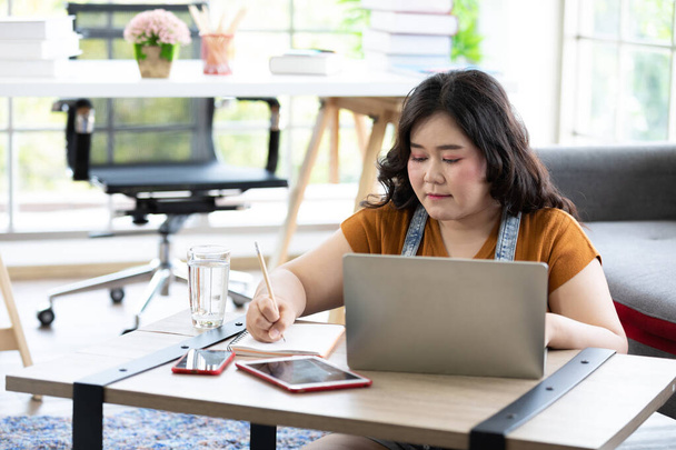 paffuto donna guardando computer portatile e la scrittura su notebook per lavoro o studio - Foto, immagini