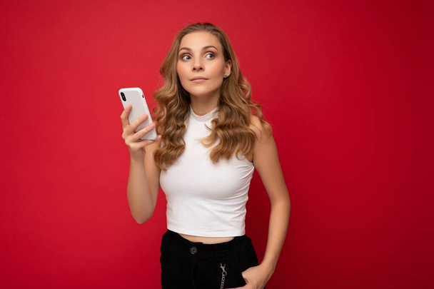gelukkig mooi jong blond vrouw dragen wit t-shirt geïsoleerd over rode achtergrond met behulp van smartphone en chatten via mobiele telefoon kijken naar de zijkant - Foto, afbeelding