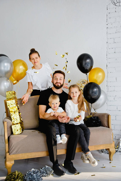 Lifestyle portrait of a mother and father with adorable children sitting on a sofa, posing for a family photo at a holiday home. Cuddle with cute little son and daughter, throw confetti - Foto, imagen