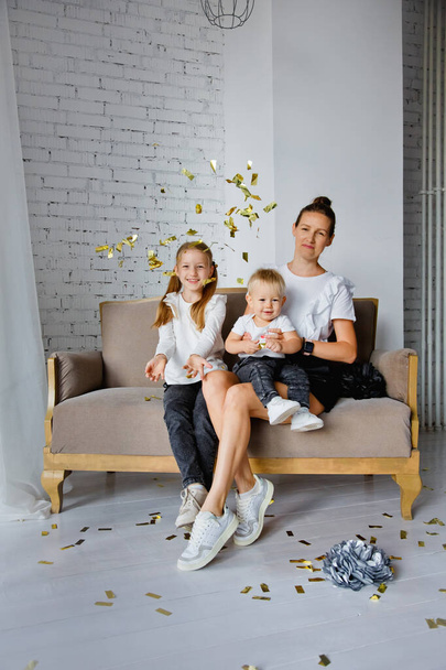 Retrato de una madre feliz con niños sentados en un sofá y posando para una foto familiar en unas vacaciones. Se acurruca con un lindo hijo pequeño y su hija mientras mira a la cámara. Lanzan confeti.. - Foto, imagen