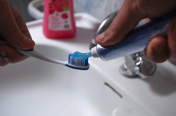 Human hands squeeze a blue toothpaste onto a blue toothbrush above the sink. - Photo, Image