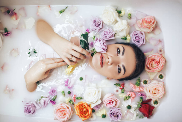 La mujer toma un baño de bienestar lleno de leche. Los brotes de rosas multicolores flotan en la superficie. Tratamientos relajantes y antienvejecimiento.. - Foto, imagen