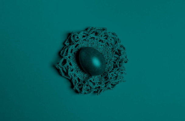 Un uovo di Pasqua giace su un nido di maglia fatto in casa. Immagine tonica nel colore di tendenza 2021 Tidewater Green. Creativo colorato minimale sfondo pasquale. Vista dall'alto. Copia spazio. - Foto, immagini