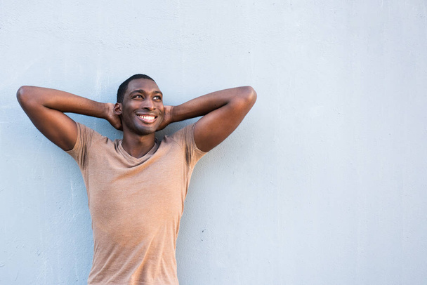 Retrato joven hombre negro sonriendo con las manos detrás de la cabeza y mirando hacia otro lado por el fondo gris - Foto, Imagen