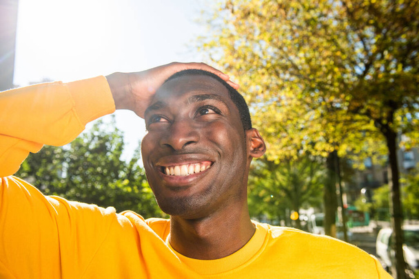 Detail portrét veselý mladý černoch usmívá s rukou na hlavě, zatímco v parku - Fotografie, Obrázek