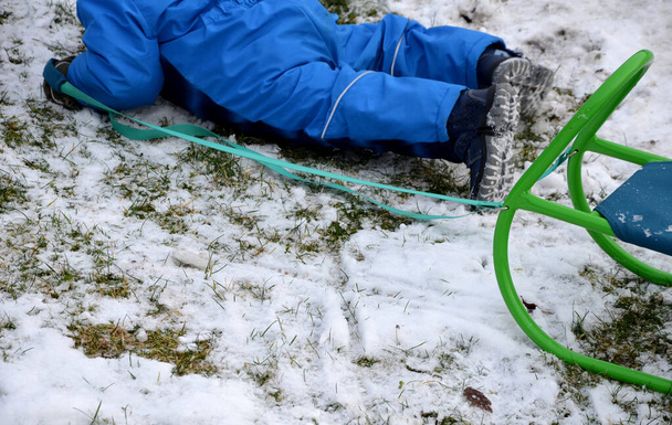 Ein kleines Kind im Winterjumpsuit versucht einen Schlitten zu ziehen und stürzt trotzdem und krabbelt kniend auf den Schneeresten herum. eine Schnur haltend und auf einem metallisch grünen Sitz sitzend, Rodeln im Garten - Foto, Bild