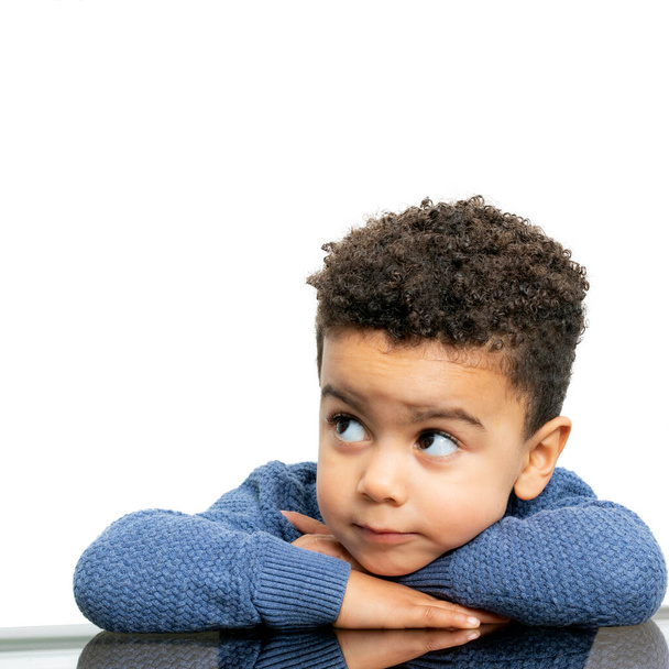 Ritratto ravvicinato di un bambino afro-americano appoggiato sulle braccia a tavola. Ragazzo che guarda da parte isolato su sfondo bianco. - Foto, immagini