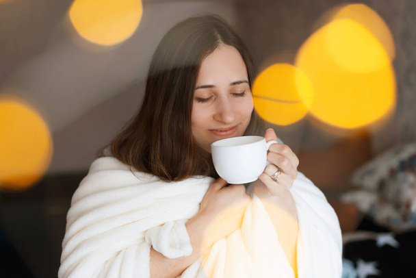 Крупным планом портрет красивой молодой женщины, нюхающей кофе или чай у окна, утреннее время - Фото, изображение