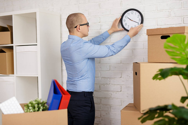 moving day concept - business man hanging clock on brick wall in new office - Photo, Image