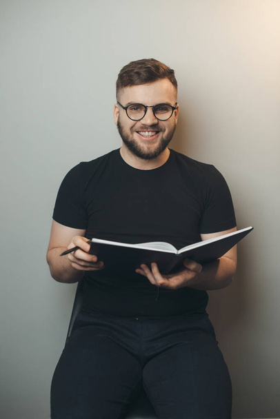 Bearded man smiling at camera on a gray studio wall holding a book and wearing glasses - Fotó, kép