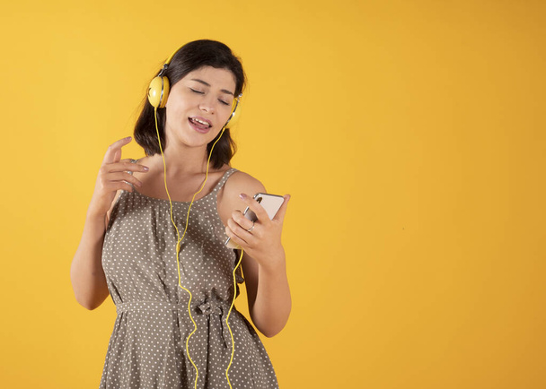 Nainen, jolla on kuulokkeet ja kännykkä, kuuntelee musiikkia ja laulaa keltaista taustaa - Valokuva, kuva