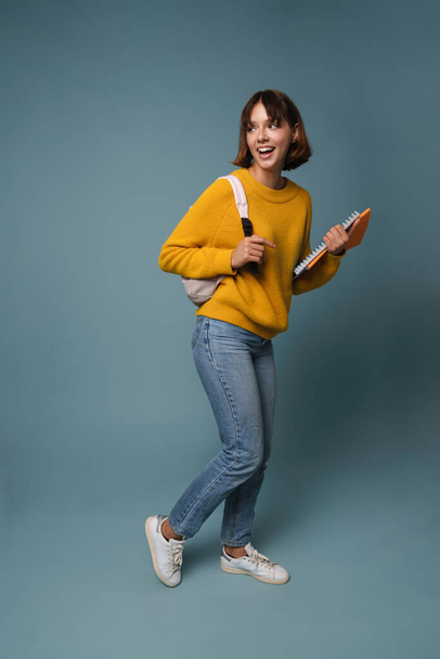 Portrait of a pretty casual student girl carrying backpack and textbooks standing isolated over blue background, full length - Фото, зображення