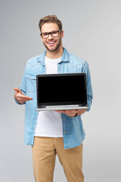 Concentrated young bearded man wearing glasses dressed in jeans shirt holding laptop isolated over grey studio background - Фото, изображение