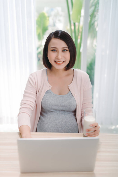 若いアジアの妊婦は自宅でコンピュータを使って仕事をし、赤ちゃんの健康のために牛乳を飲む. - 写真・画像