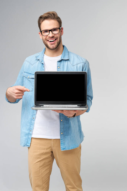 Concentrated young bearded man wearing glasses dressed in jeans shirt holding laptop isolated over grey studio background - Foto, immagini