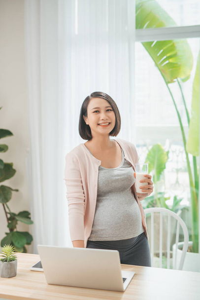 Fiatal szépség boldog ázsiai terhes nő kezében pohár tej segítségével számítógép otthon. - Fotó, kép