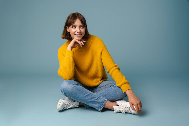 Portrait of smiling young woman resting while sitting on a floor with legs crossed isolated over blue background - Fotoğraf, Görsel