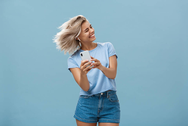 Great vibes carrying me away. Carefree joyful attractive young blonde female in stylish outfit waving head and smiling broadly listening music in wireless earbuds and holding smartphone over blue wall - Photo, Image