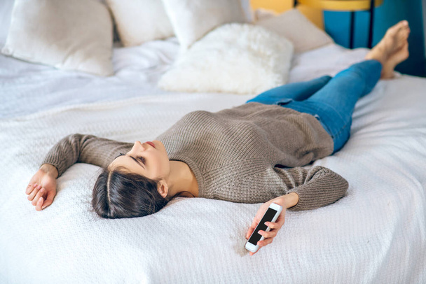 Dark-haired young woman lying on bed with a phone in her hand and looking unhappy - 写真・画像