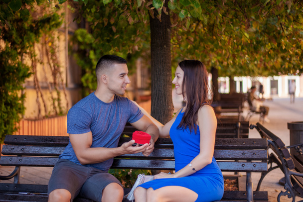Una pareja amorosa sentada en un banco un hombre le da a una chica una caja en forma de corazón. El día del amor. - Foto, imagen