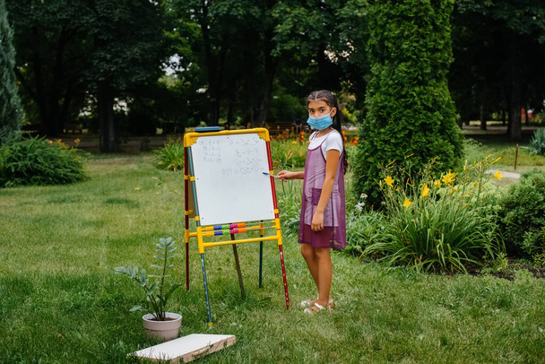 Μια μαθήτρια με μάσκα στέκεται και γράφει μαθήματα στον πίνακα. Επιστροφή στο σχολείο, μάθηση κατά τη διάρκεια της πανδημίας - Φωτογραφία, εικόνα