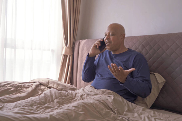 Счастливый улыбающийся бизнес, черный мужчина, афроамериканцы разговаривают по смартфону на кровати в спальне дома. Концепция устройства в технике с раннего утра. - Фото, изображение