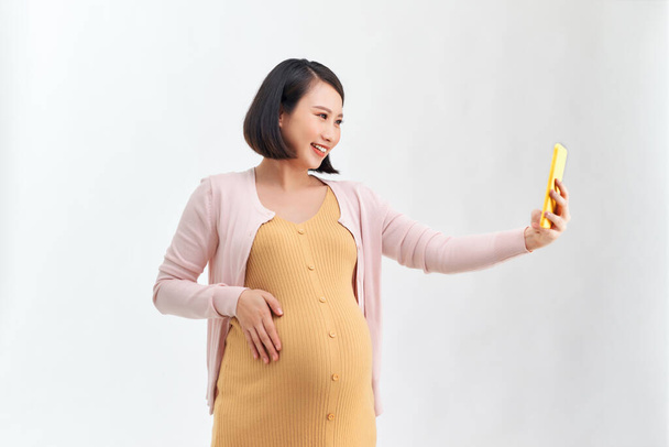 щаслива вагітна жінка зі смартфоном бере селфі на білому тлі
 - Фото, зображення