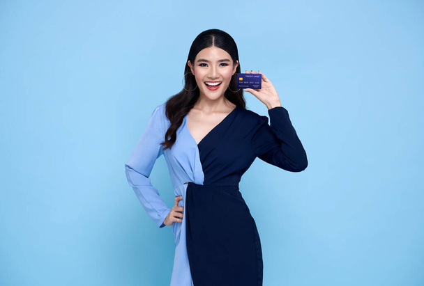 Happy beautiful Asian shopaholic women wearing blue dress showing credit card in hand isolated on blue background. - Photo, Image